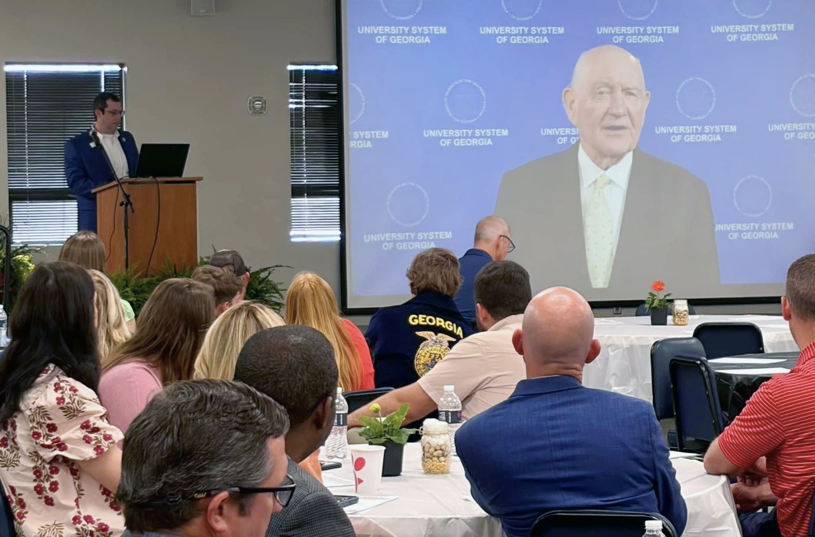 a group of people watching a man speak on a screen